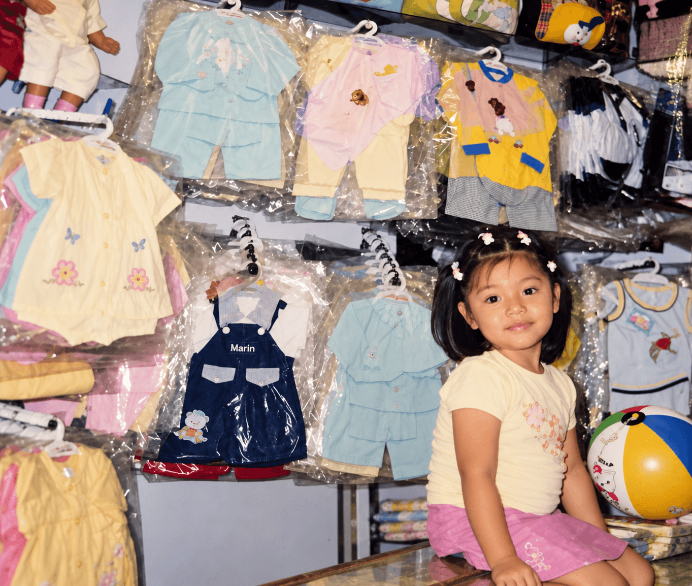 Sayuri de niña en la tienda de ropa del negocio familiar, rodeada de prendas colgadas